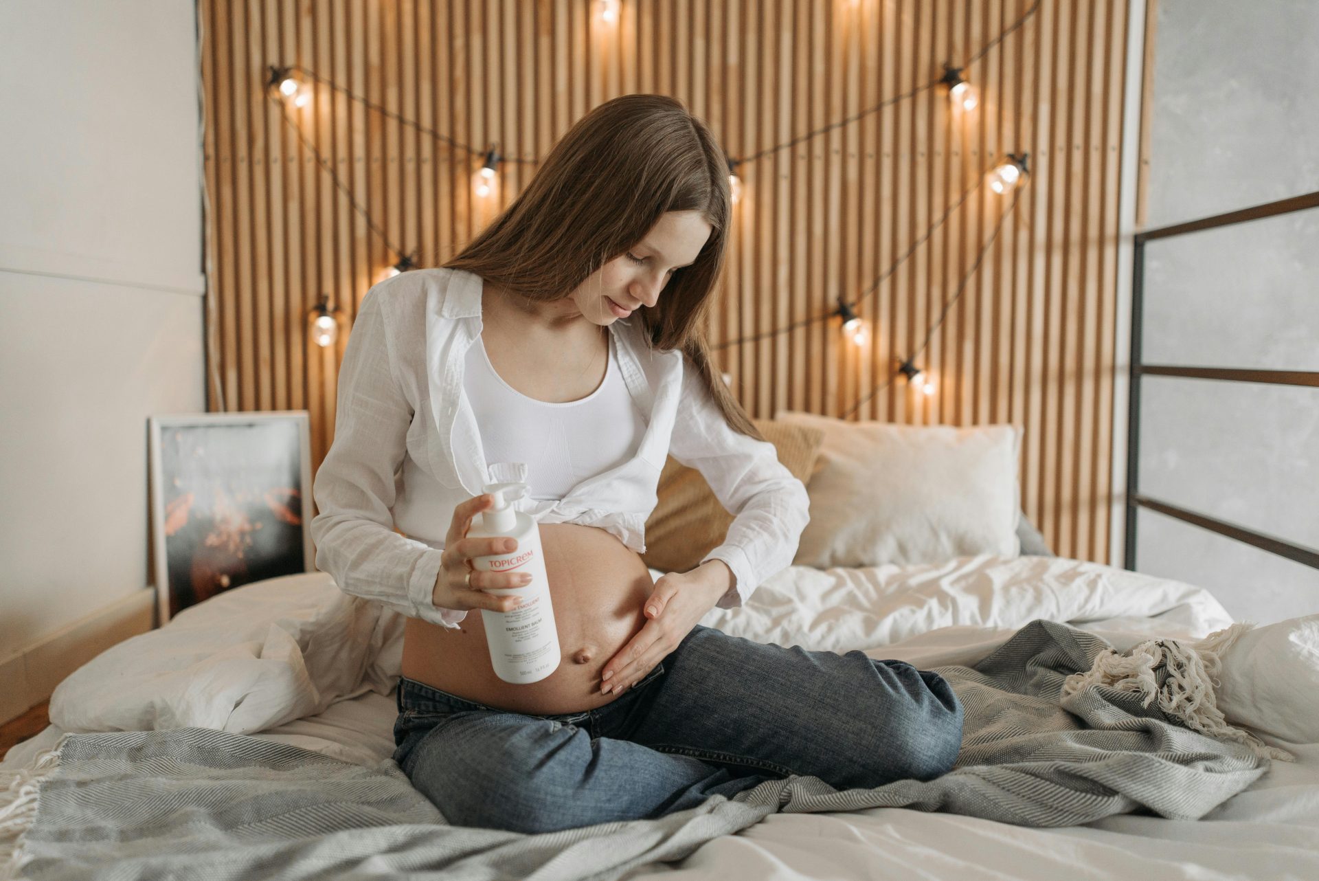 Eine werdende Mutter pflegt ihren Bauch mit Lotion in gemütlicher Schlafzimmeratmosphäre.
