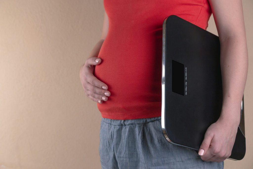 Schwangere Frau berührt ihren Bauch und trägt eine digitale Badezimmerwaage.